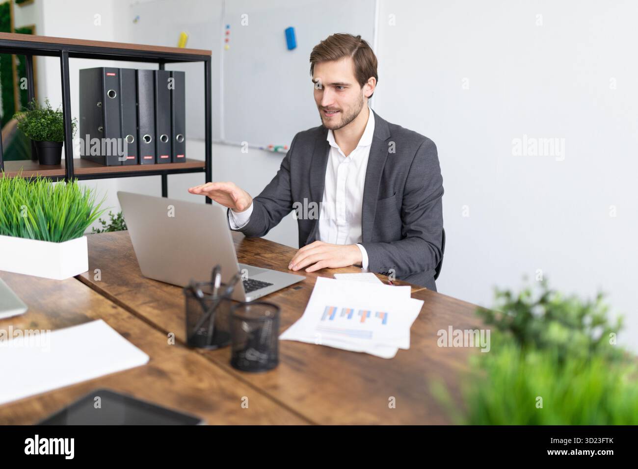 Uomo d'affari che tiene un seminario online seduto a un tavolo di fronte a un portatile con documenti e documenti in ufficio Foto Stock