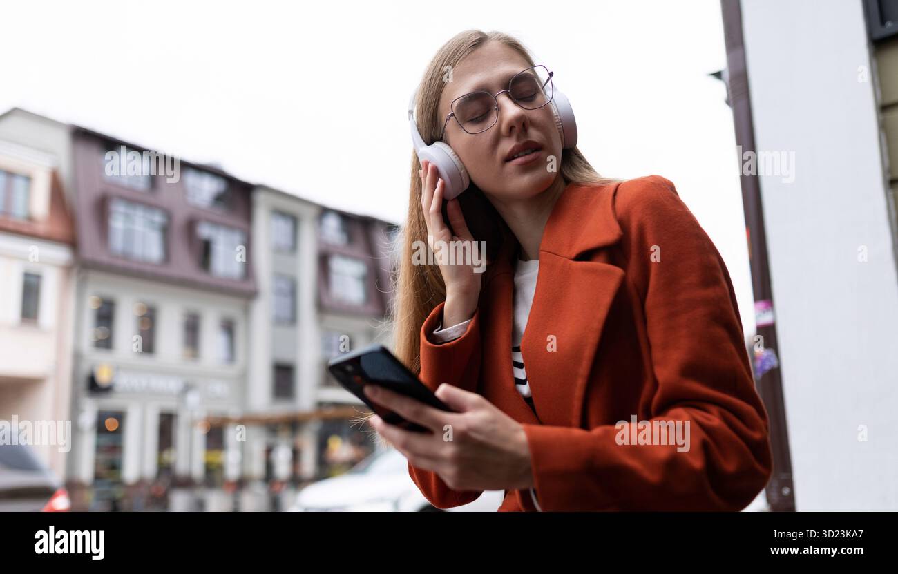 La giovane donna bionda con un telefono cellulare in mano ascolta la musica con le cuffie sullo sfondo del paesaggio urbano Foto Stock