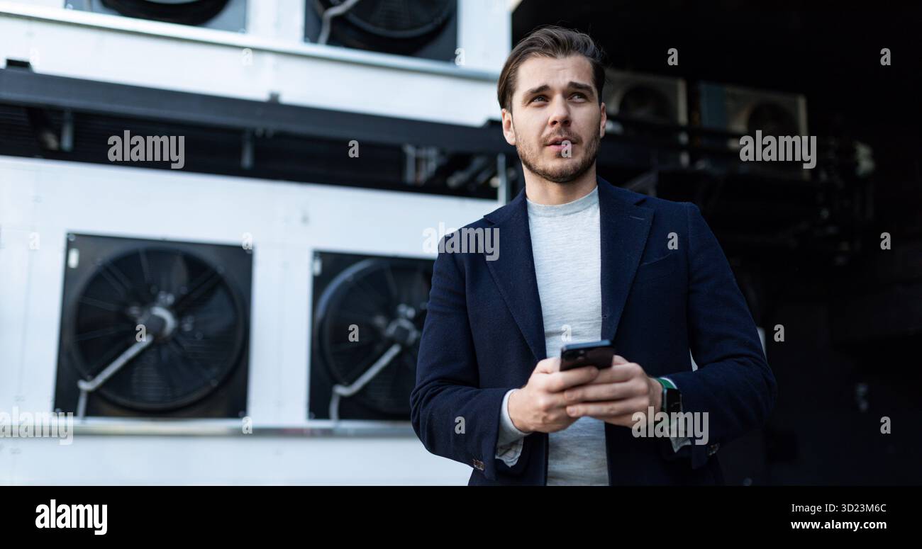 Ingegnere installatore di condizionatori d'aria sullo sfondo di apparecchiature di refrigerazione industriali con un telefono cellulare in mano Foto Stock