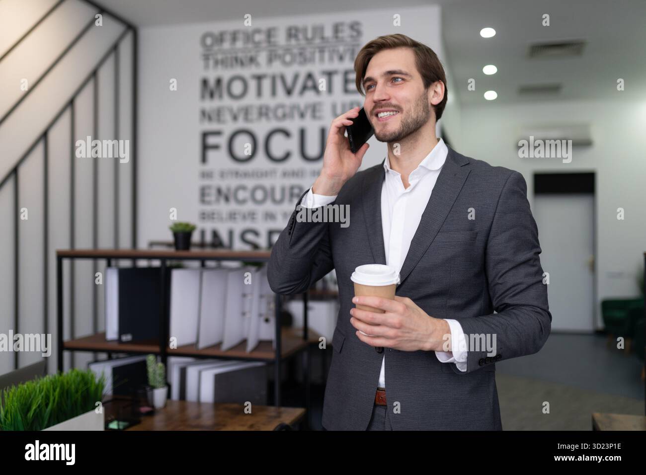 Uomo d'affari in tuta formale che parla al telefono con una tazza di caffè tra le mani in ufficio Foto Stock