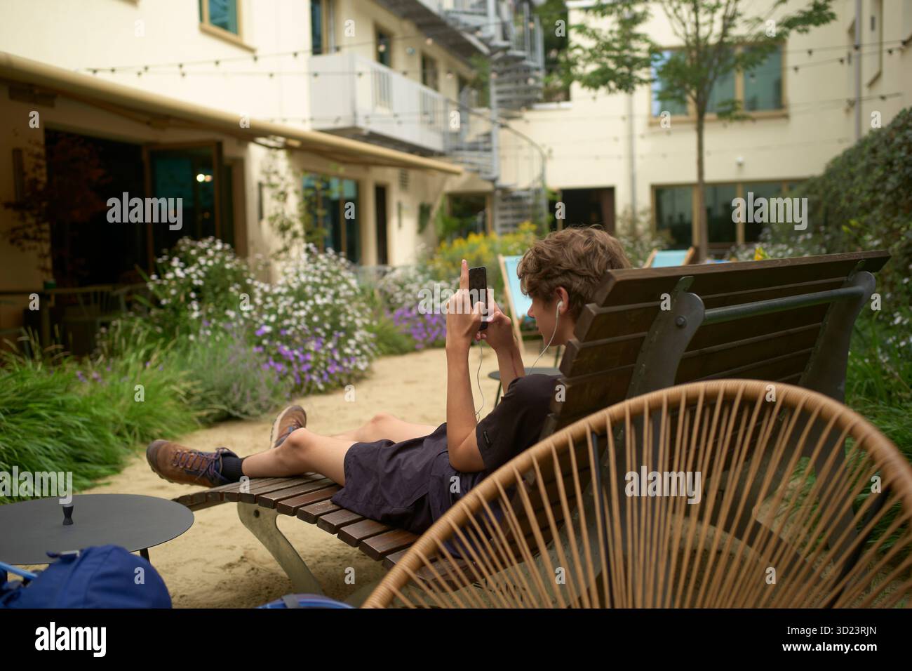Un giovane adulto rilassato che usa il telefono mentre si rilassa su una sedia di legno in un cortile con giardino. Monaco, Germania Foto Stock