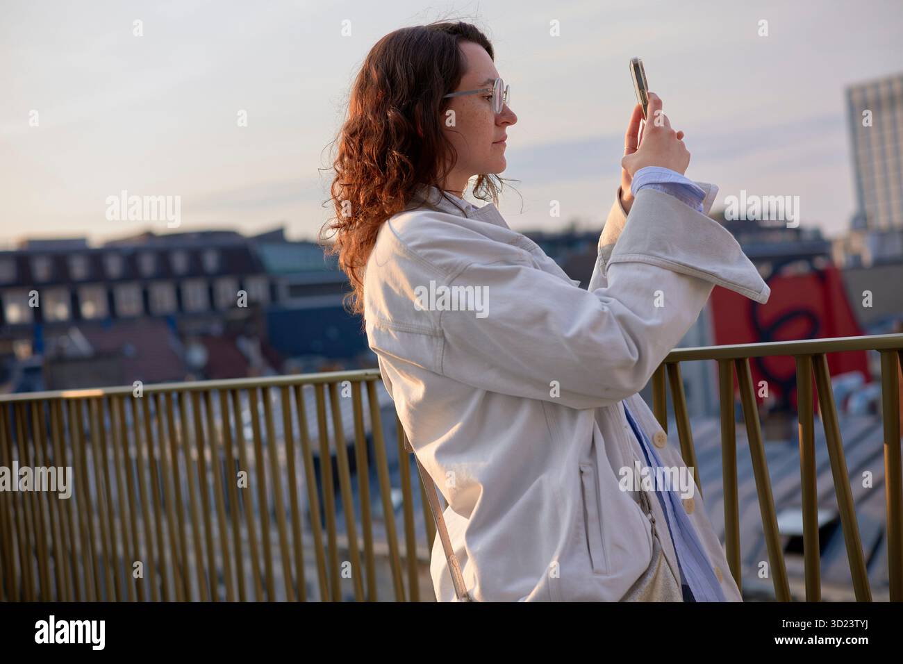 Una donna con un cappotto bianco scatta una foto su un balcone con un paesaggio urbano sullo sfondo. Bruxelles, Belgio Foto Stock