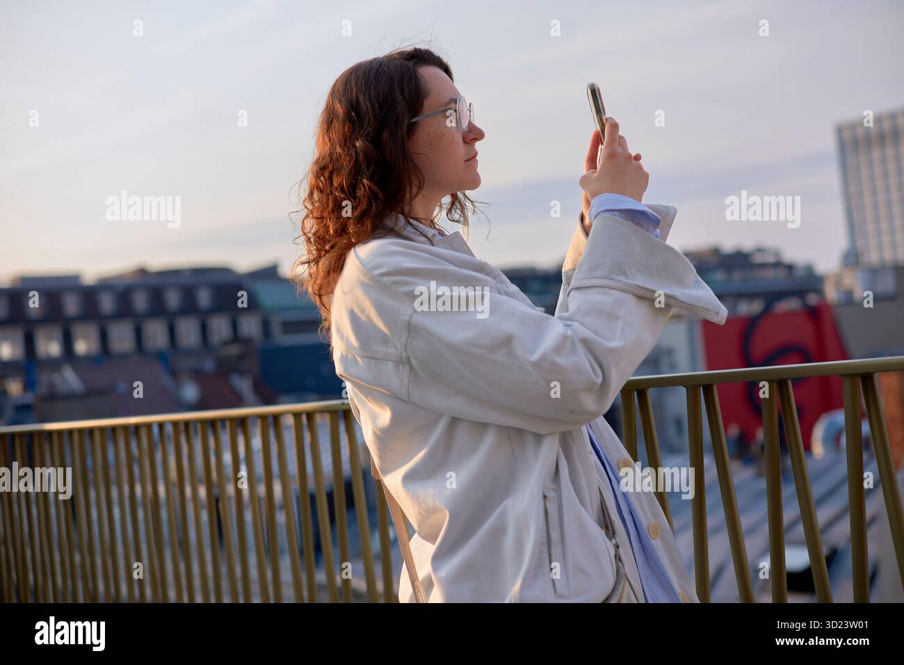 Una donna con un cappotto bianco scatta una foto su un balcone con un paesaggio urbano sullo sfondo. Bruxelles, Belgio Foto Stock