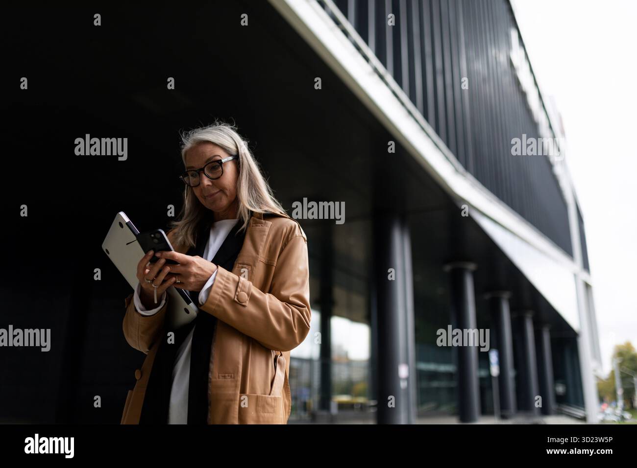 Una donna d'affari anziana di mezza età guarda il telefono con un portatile tra le mani accanto al centro ufficio, lavora fuori dal Foto Stock