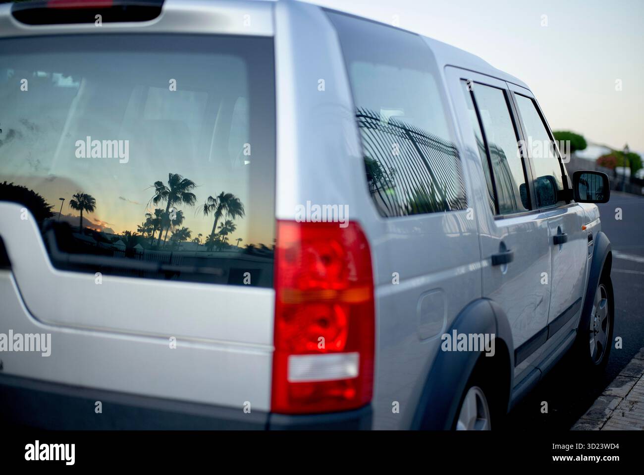 Il SUV Silver parcheggiato sulla strada riflette le palme nel finestrino posteriore al tramonto. Lanzarote, Spagna Foto Stock