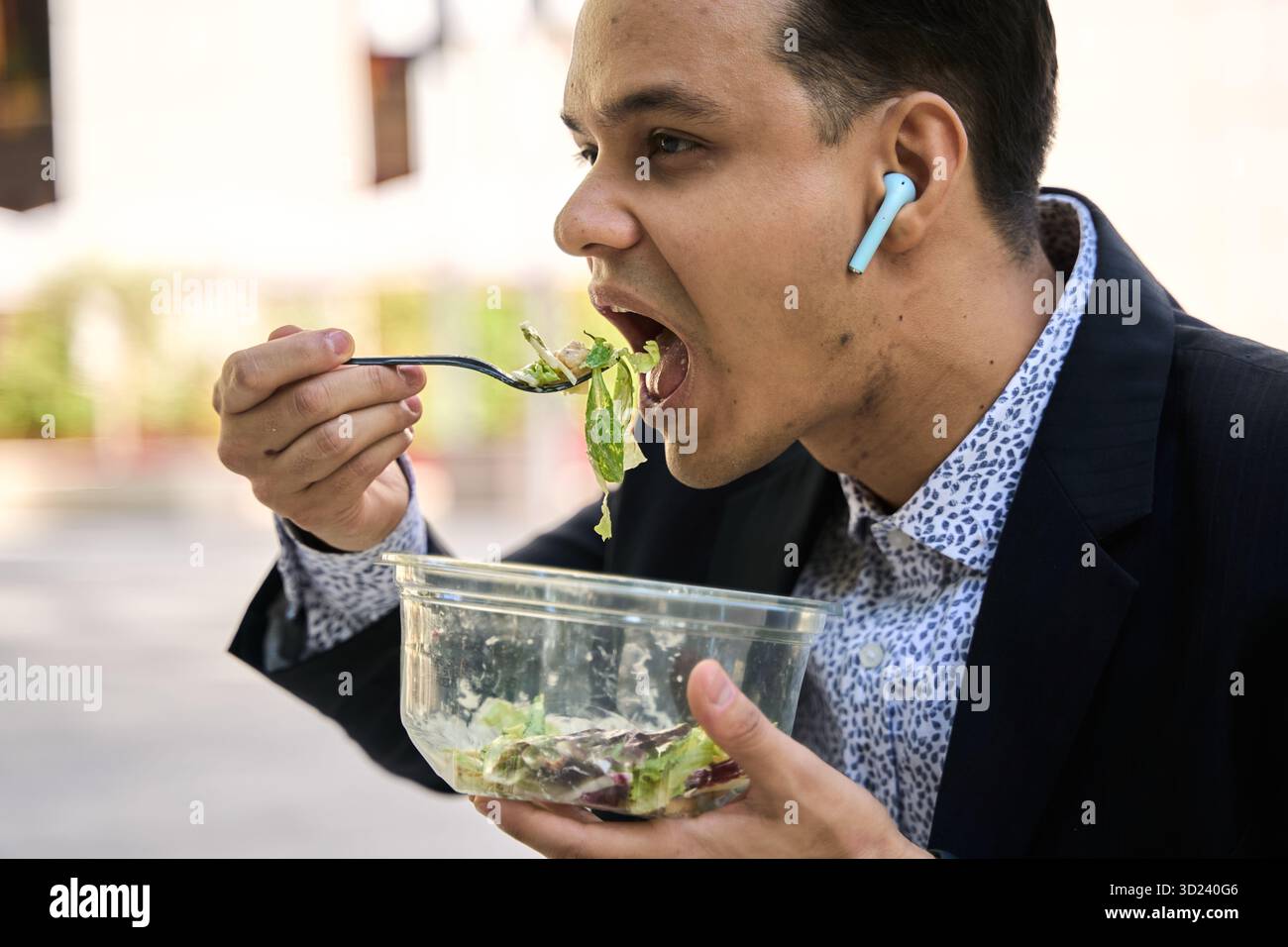 Giovane uomo latino in costume da lavoro che gusta una sana insalata fresca all'aperto durante la pausa pranzo, indossando auricolari wireless Foto Stock