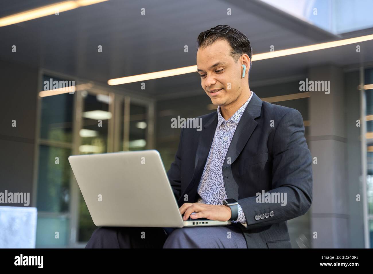 Uomo d'affari latino in tuta che usa un computer portatile con auricolari wireless, lavora a distanza all'esterno di un moderno edificio in vetro Foto Stock