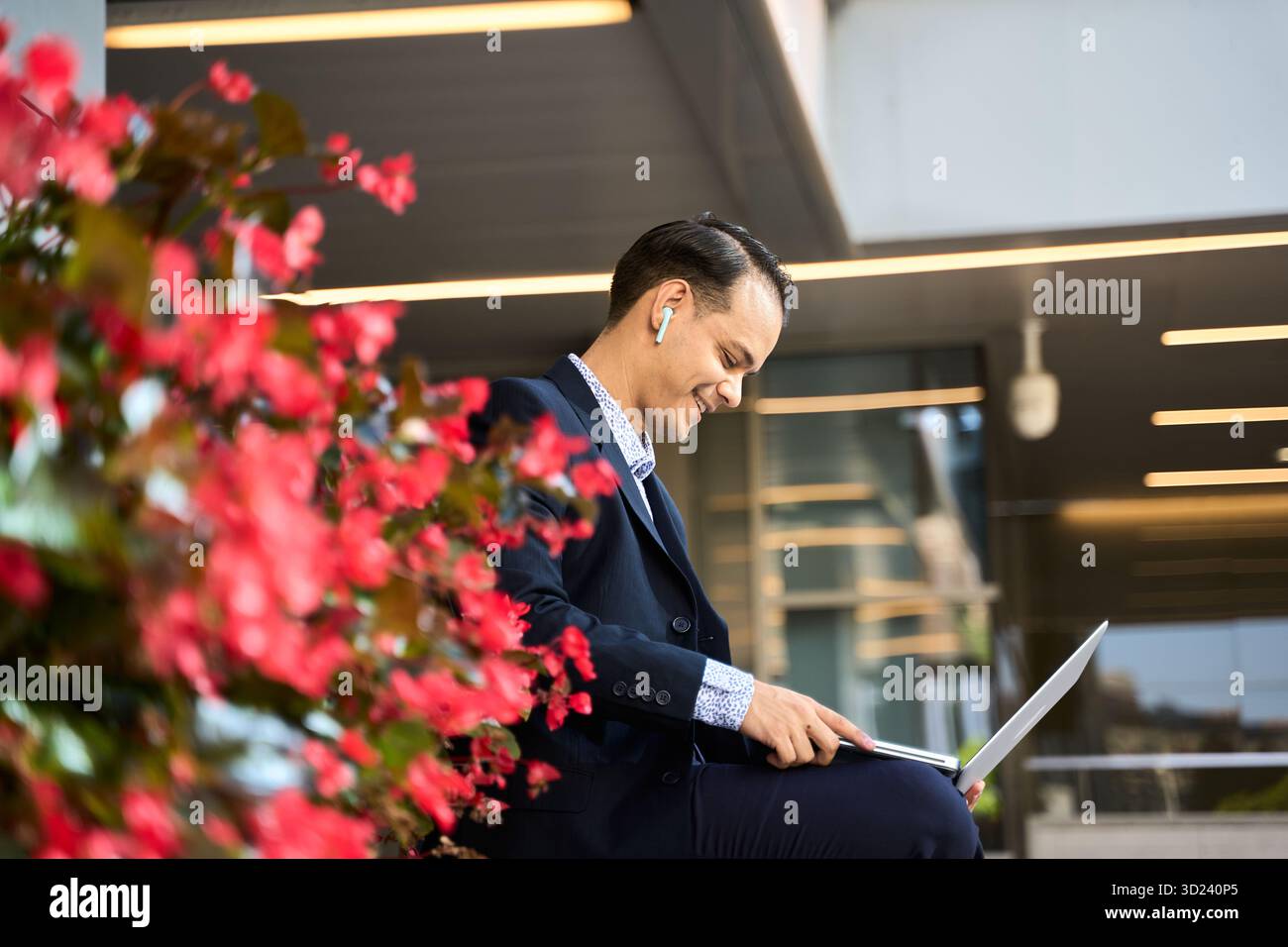 Un uomo d'affari latino sorridente che usa laptop e auricolari all'aperto, seduto accanto a fiori vivaci in un moderno ambiente urbano Foto Stock
