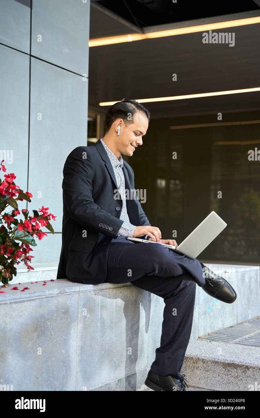 Uomo d'affari latino in tuta formale che lavora all'aperto su un notebook, sorridendo con gli auricolari wireless, simboleggiando il lavoro da remoto e la produttività Foto Stock
