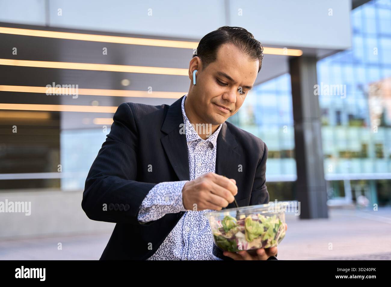 Uomo d'affari latino in costume che gusta un'insalata fresca all'aperto, simboleggia uno stile di vita sano e un moderno equilibrio aziendale Foto Stock
