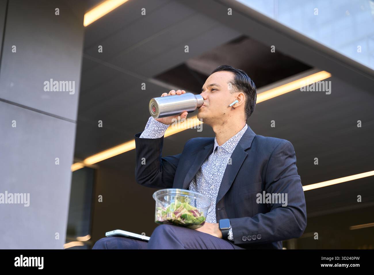 Uomo d'affari latino in abito formale che gusta un'insalata e una bevanda durante la pausa pranzo all'aperto in un ambiente urbano moderno Foto Stock