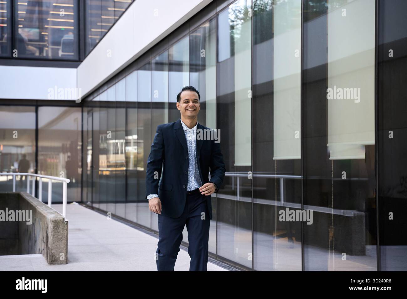 Uomo d'affari latino sorridente in tuta formale che cammina all'aperto vicino a un moderno edificio di uffici in vetro, mostrando successo e fiducia Foto Stock