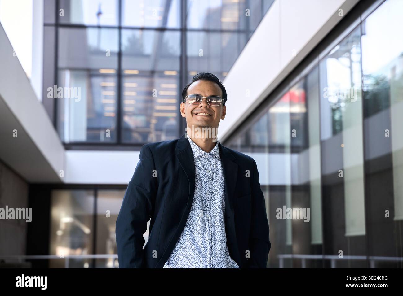 Uomo d'affari latino in abiti formali sorridente con fiducia mentre si trova in un moderno corridoio di un edificio di vetro Foto Stock