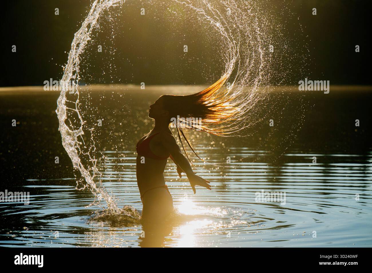 Una donna dalla silhouette ribalta i capelli, creando spruzzi d'acqua al tramonto. Wildcat Lake Park, Washington, Stati Uniti Foto Stock