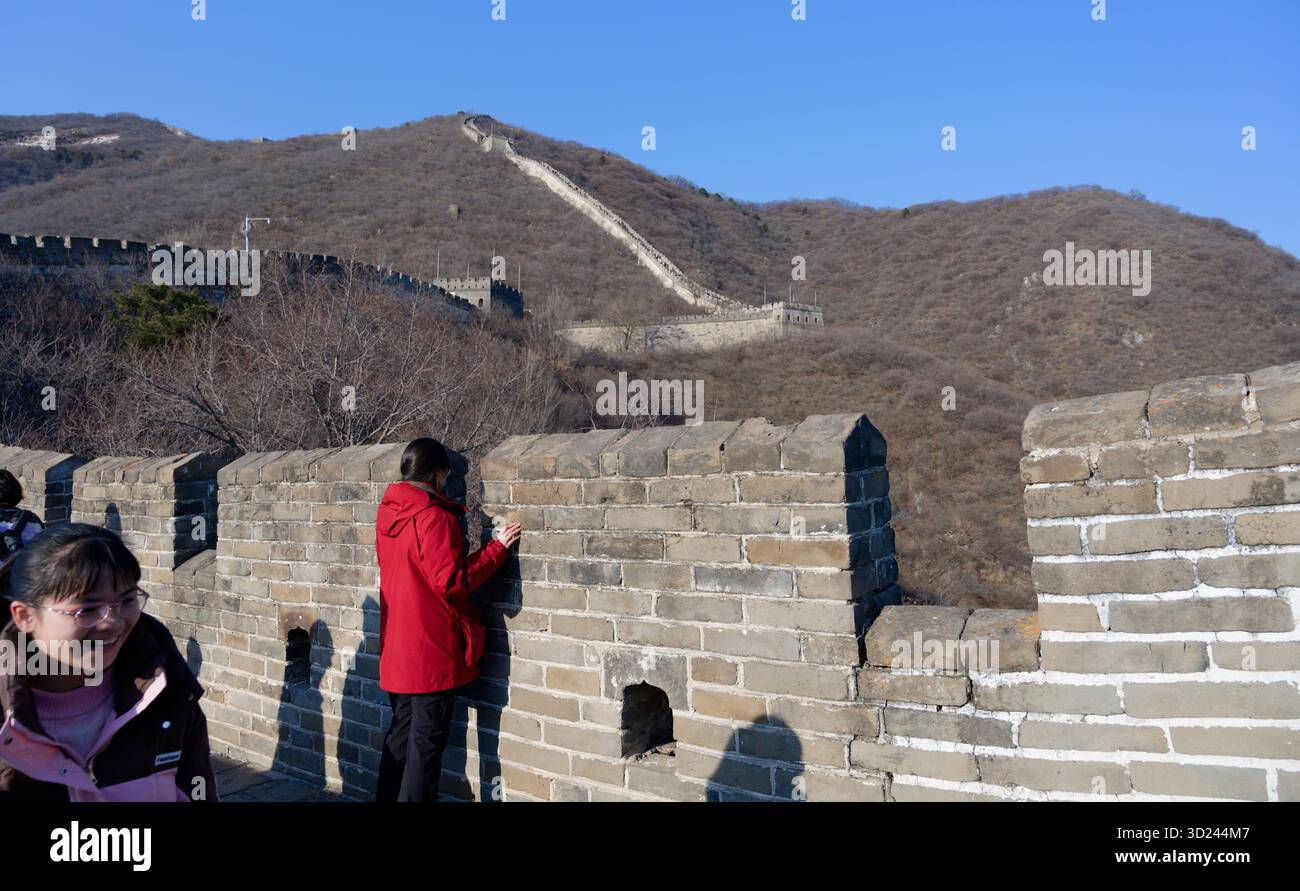 Donna con una giacca rossa guarda attraverso una delle merlature della grande Muraglia Cinese Foto Stock