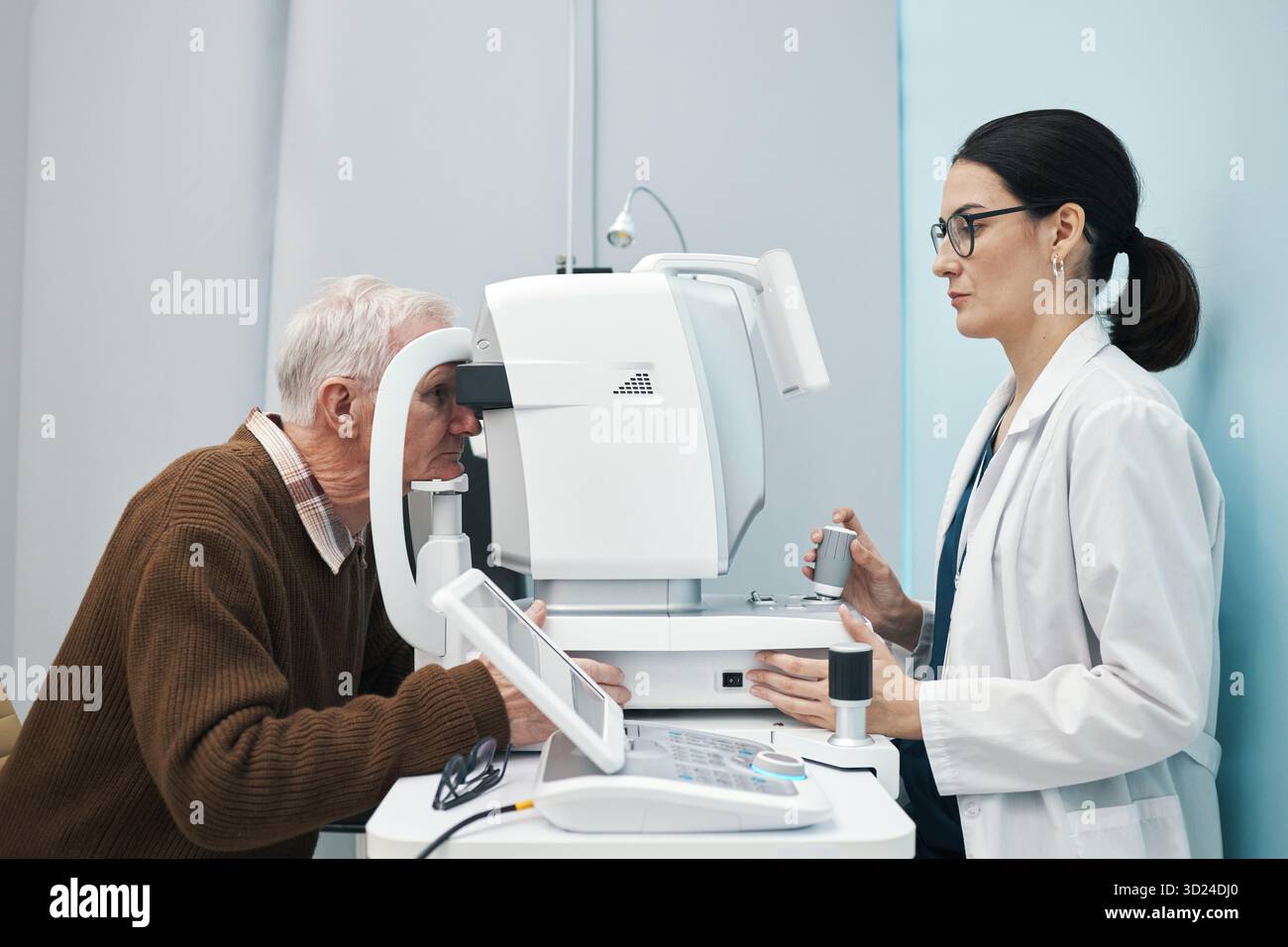 Uomo caucasico anziano sottoposto a esame oculistico con dispositivo oftalmico operato da giovane medico donna caucasica adulta, entrambi concentrati sulla procedura in ambiente clinico Foto Stock