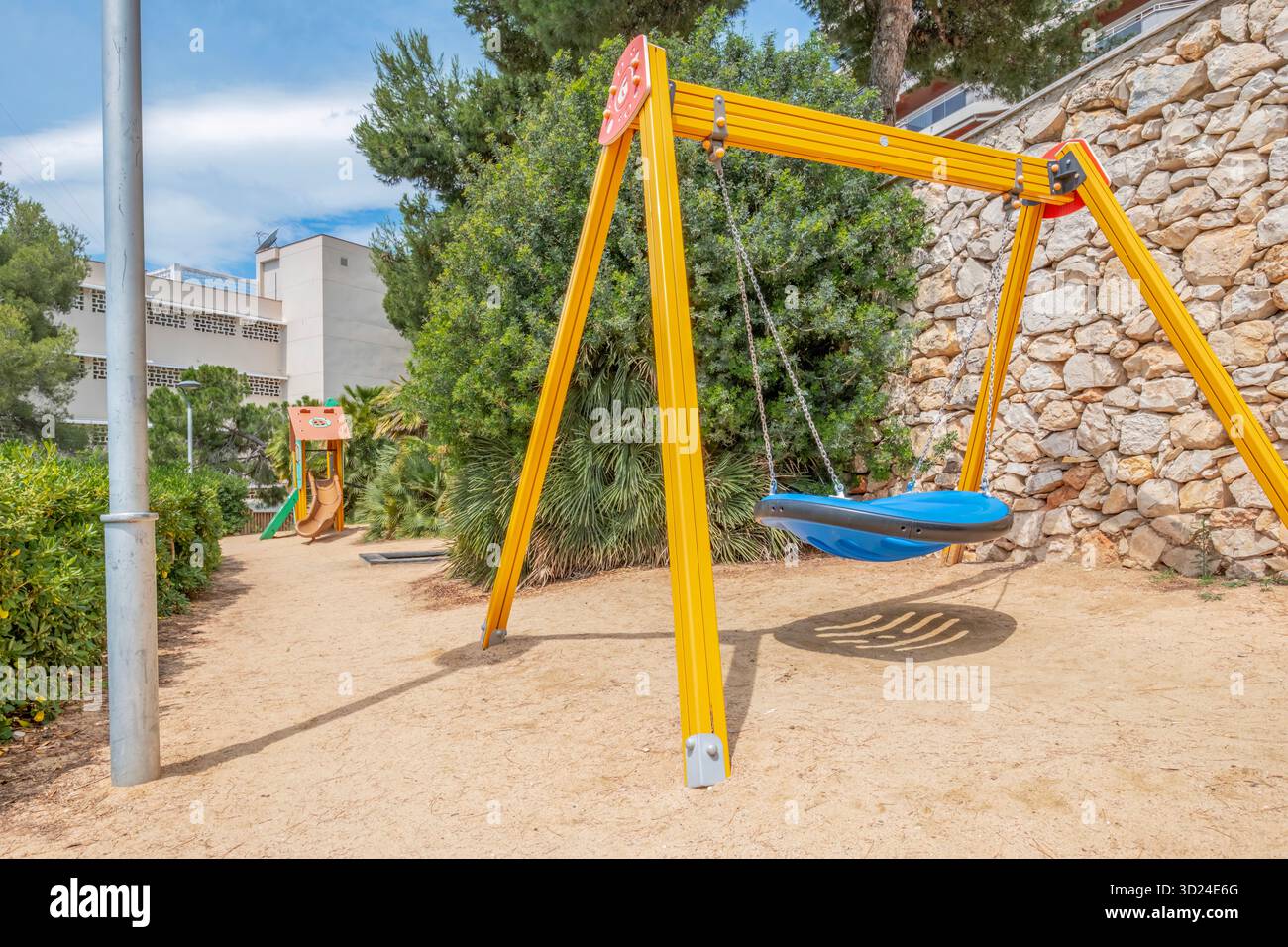 Svuota il cesto blu su un'altalena gialla situata in un parco giochi soleggiato e recintato con terreno sabbioso, cespugli verdi e muri di pietra Foto Stock