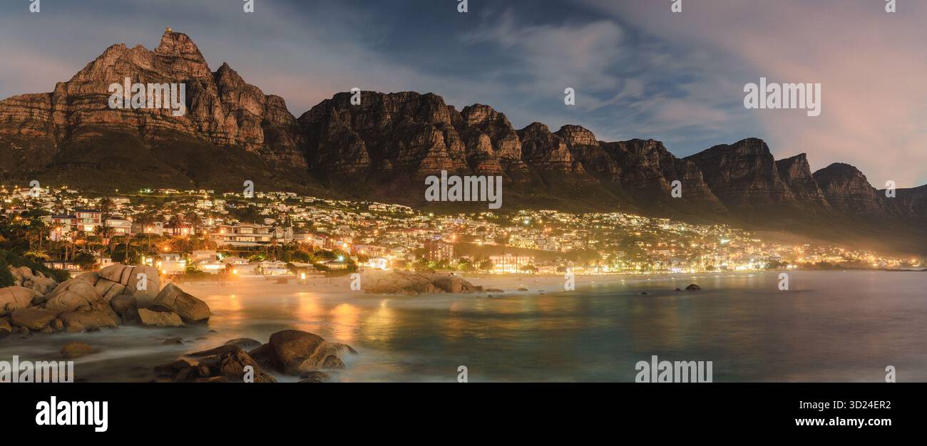 Vista delle luci della città che brillano come gioielli sparsi sotto l'imponente profilo di una aspra catena montuosa, riflessa nelle acque tranquille, Foto Stock