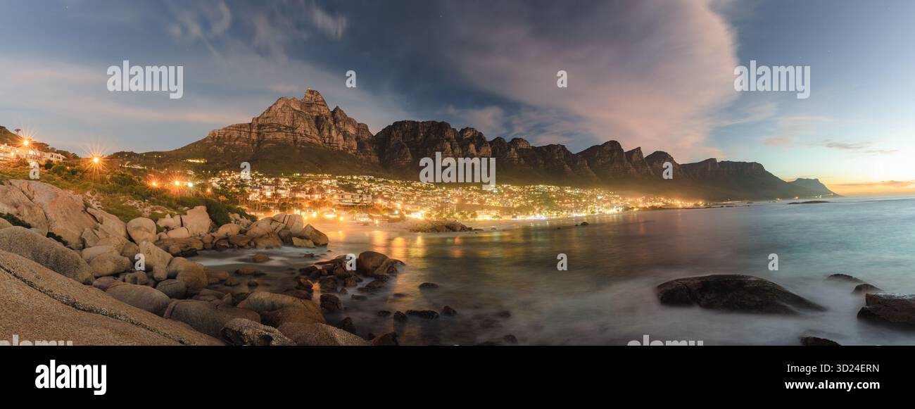 La vista delle luci della città brilla lungo la costa, riflettendo sulle acque tranquille sotto l'osservazione delle maestose montagne, città del Capo, Capo Occidentale, Foto Stock