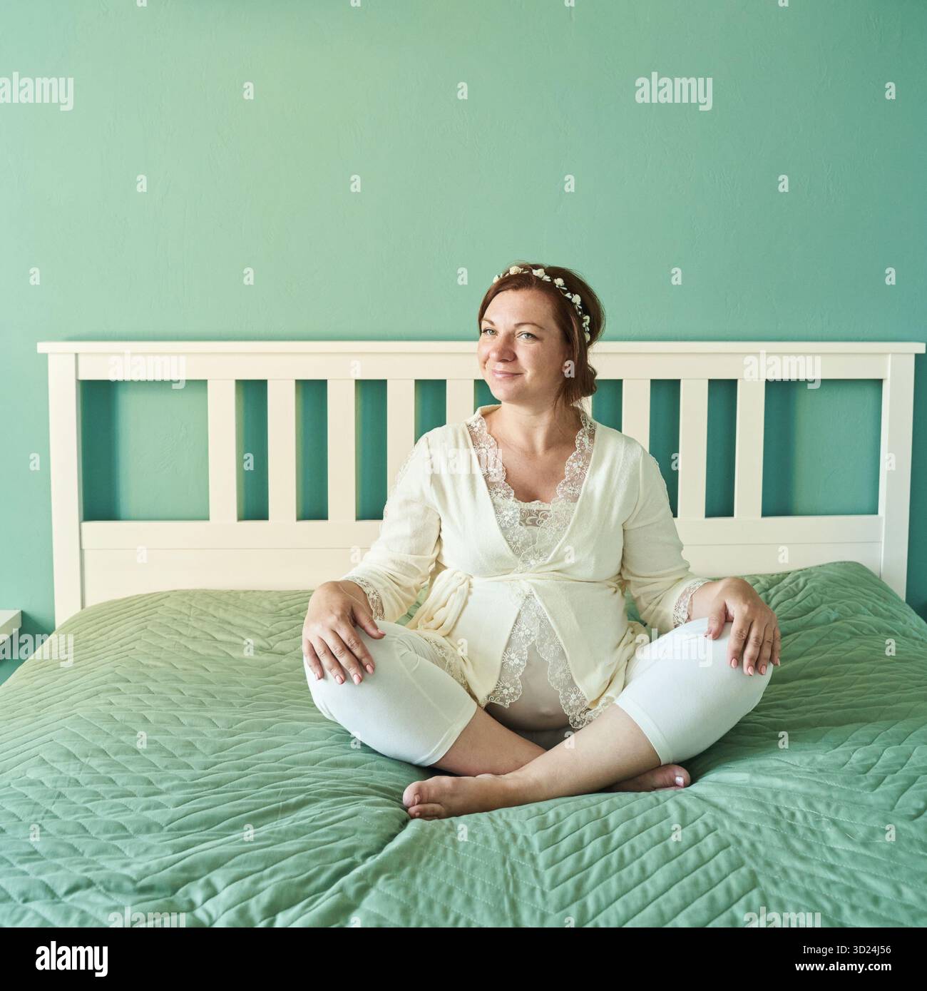 Donna caucasica adulta seduta comodamente sul letto con biancheria da letto verde chiaro Foto Stock