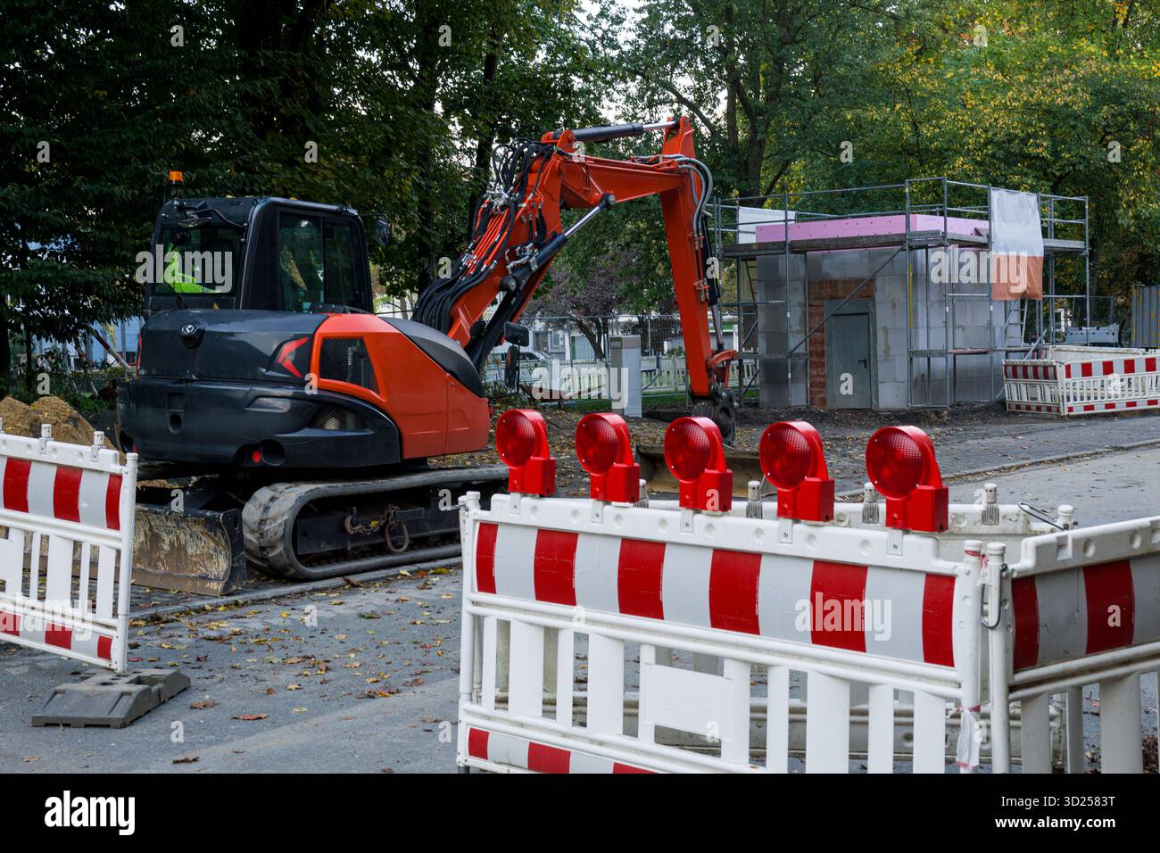 Cantiere stradale con barriere di sicurezza rosse e bianche e escavatore in funzione vicino all'area residenziale. Foto Stock