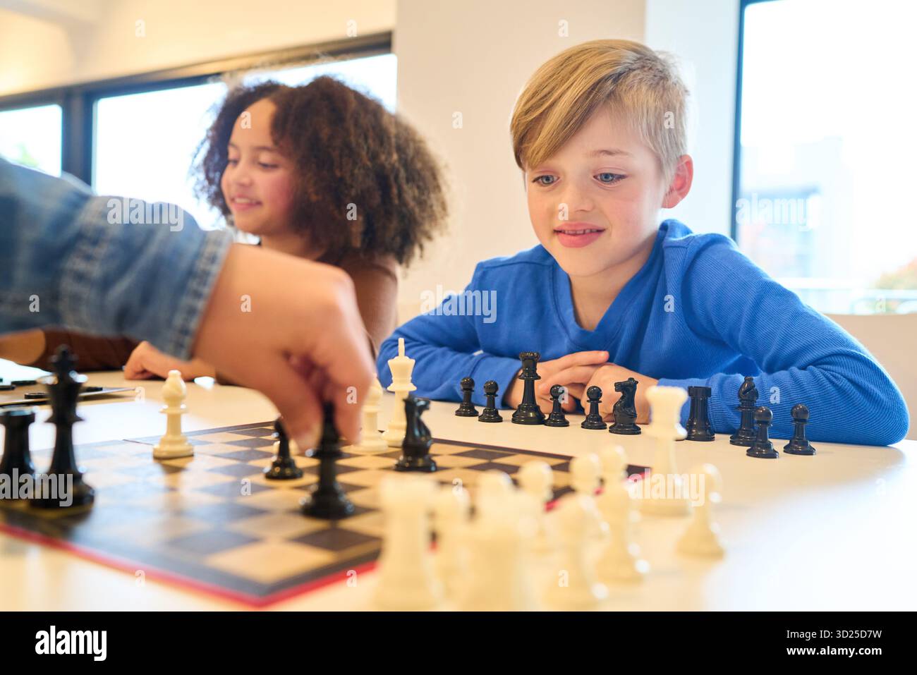 Due bambini siedono uno di fronte all'altro a una scacchiera in uno spazio luminoso e accogliente, incentrato sul gioco. La scena enfatizza la concentrazione, cooperat Foto Stock
