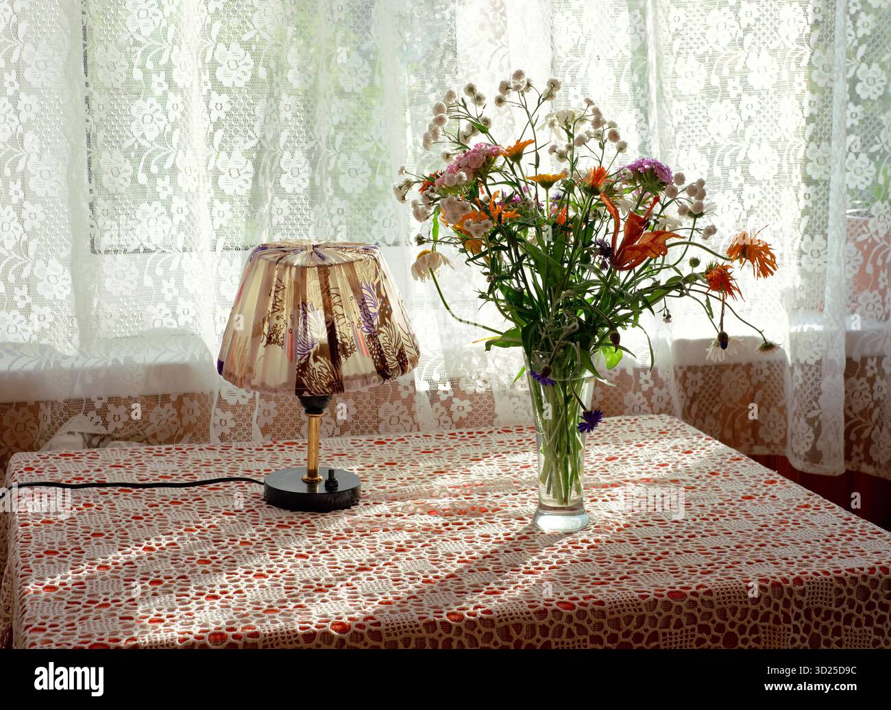 Vaso in vetro che tiene fiori selvatici assortiti in piedi sul tavolo accanto a una piccola lampada decorativa, la luce del sole che filtra attraverso le tende in pizzo sullo sfondo creando intricati motivi d'ombra Foto Stock