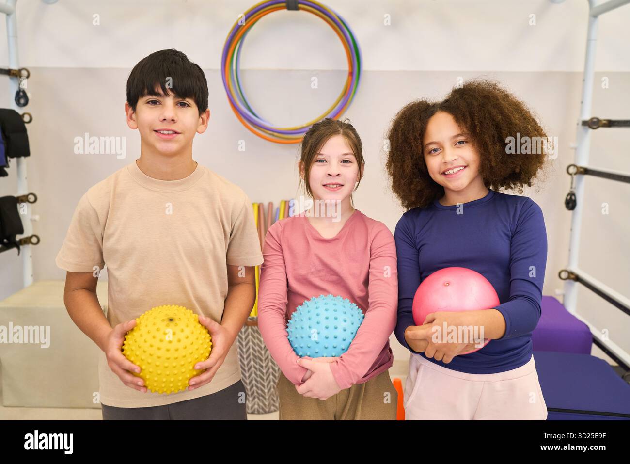 Tre bambini sorridenti stanno insieme in una sala riabilitativa, ognuno con una palla colorata. L'impostazione prevede cerchi e tappetini morbidi, alti Foto Stock