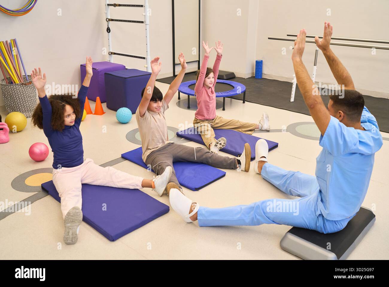 Una sessione di terapia pediatrica in una palestra di riabilitazione dove i bambini e un medico eseguono esercizi di ginnastica seduti e di equilibrio su tappeti blu utilizzando suppor Foto Stock