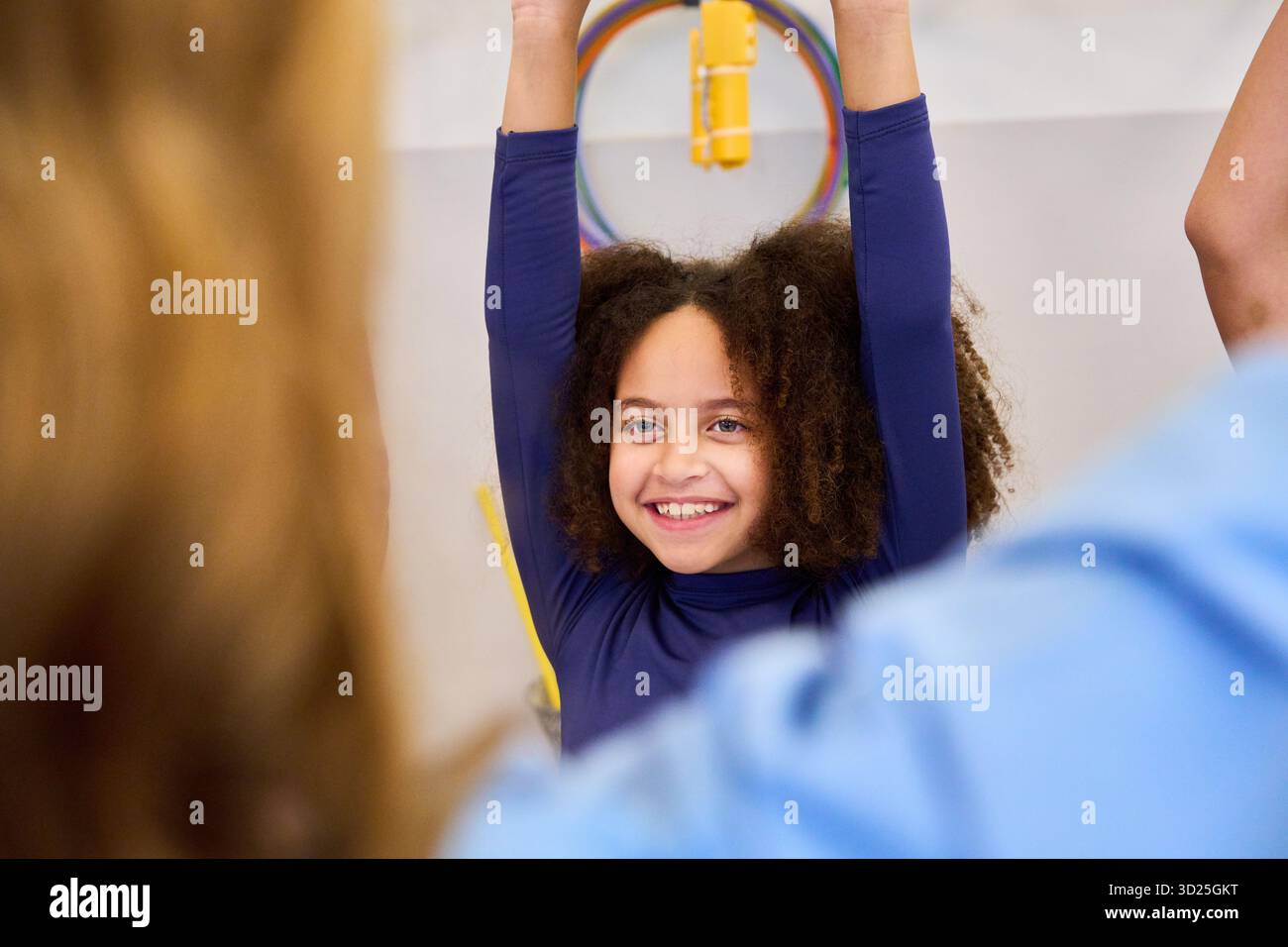 Una ragazza sorridente con la parte superiore blu a maniche lunghe allunga le braccia durante una sessione di riabilitazione pediatrica, guidata da un terapeuta in un ambiente clinico. Foto Stock