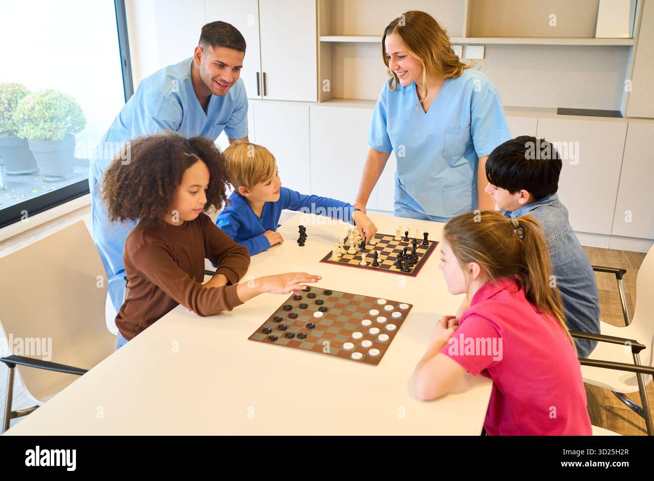 Brillante scena clinica pediatrica in cui i terapisti in scrub blu guidano i bambini mentre giocano a scacchi, aumentando la concentrazione, le abilità sociali e il coordinamento in A. Foto Stock