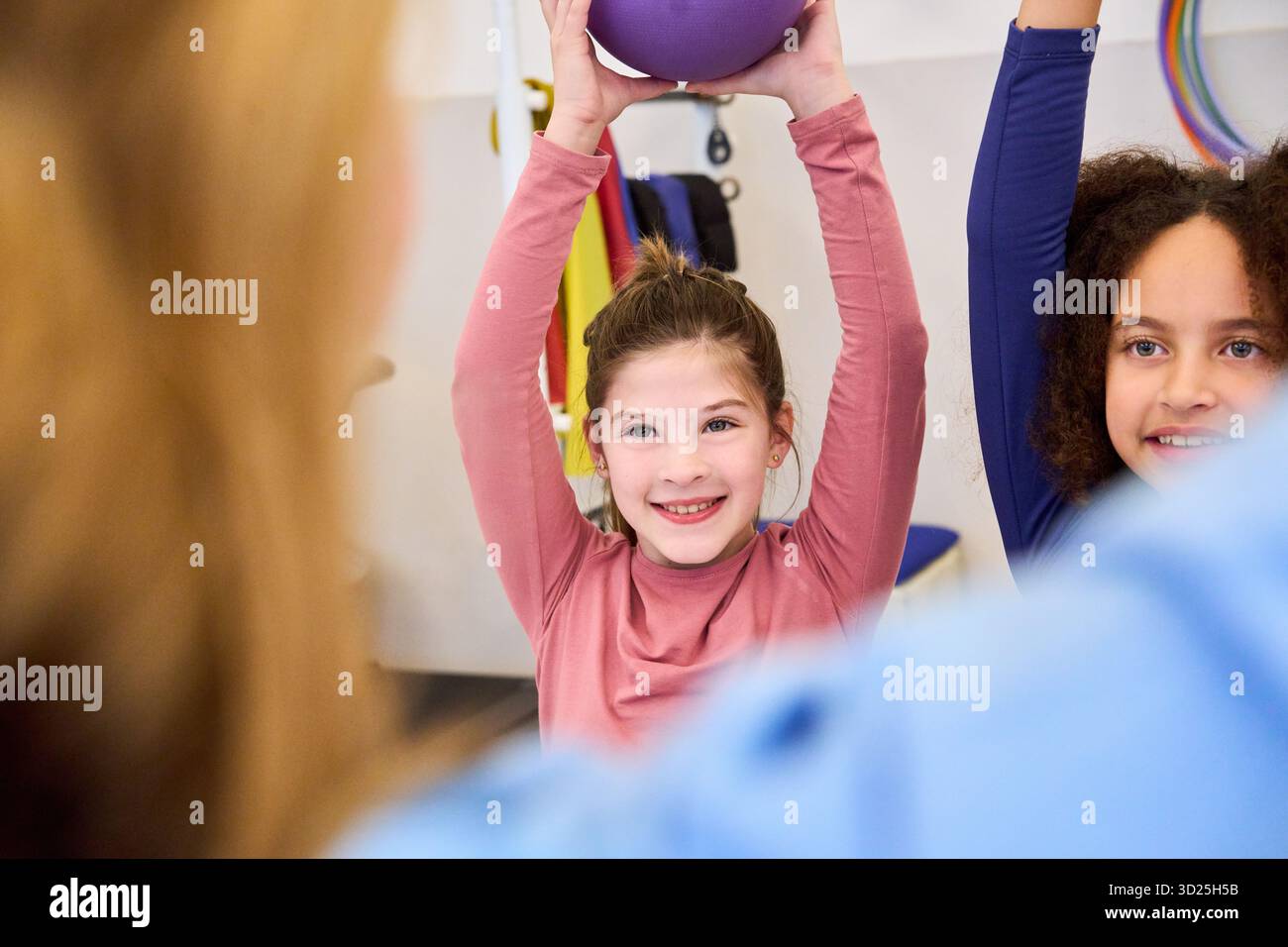 Un bambino sorridente in cima rosa partecipa a un'attività di fisioterapia di gruppo, sollevando una palla viola sopra la testa. In un ambiente di riabilitazione luminoso, peer Foto Stock