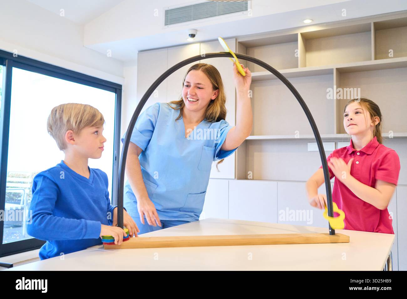 Un terapista amichevole conduce due bambini in un ambiente di riabilitazione pediatrica, guidando l'equilibrio e le attività di coordinamento con un cerchio e un professionista leggero Foto Stock