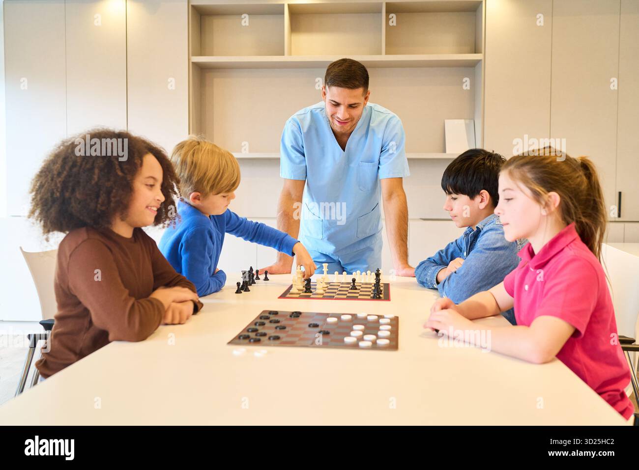 Un medico cordiale guida un gruppo di bambini in una dolce attività riabilitativa in una clinica luminosa. Esercitano le capacità motorie e la risoluzione dei problemi Foto Stock