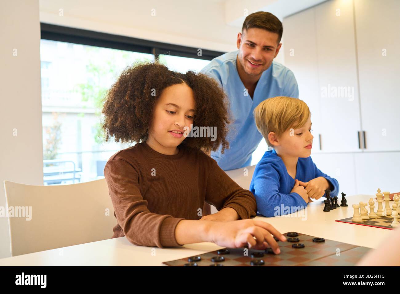 Un terapeuta amichevole supervisiona due bambini mentre si impegnano in un'attività da tavolo, combinando abilità motorie con giochi cognitivi durante un pediat Foto Stock