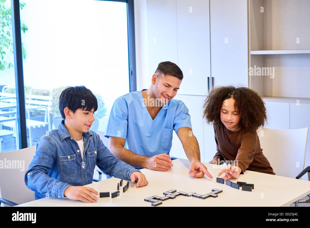 Un terapeuta in scrub blu conduce due bambini in attività motorie e di equilibrio utilizzando blocchi in una clinica soleggiata e accogliente. Assistenza mirata e di supporto Foto Stock