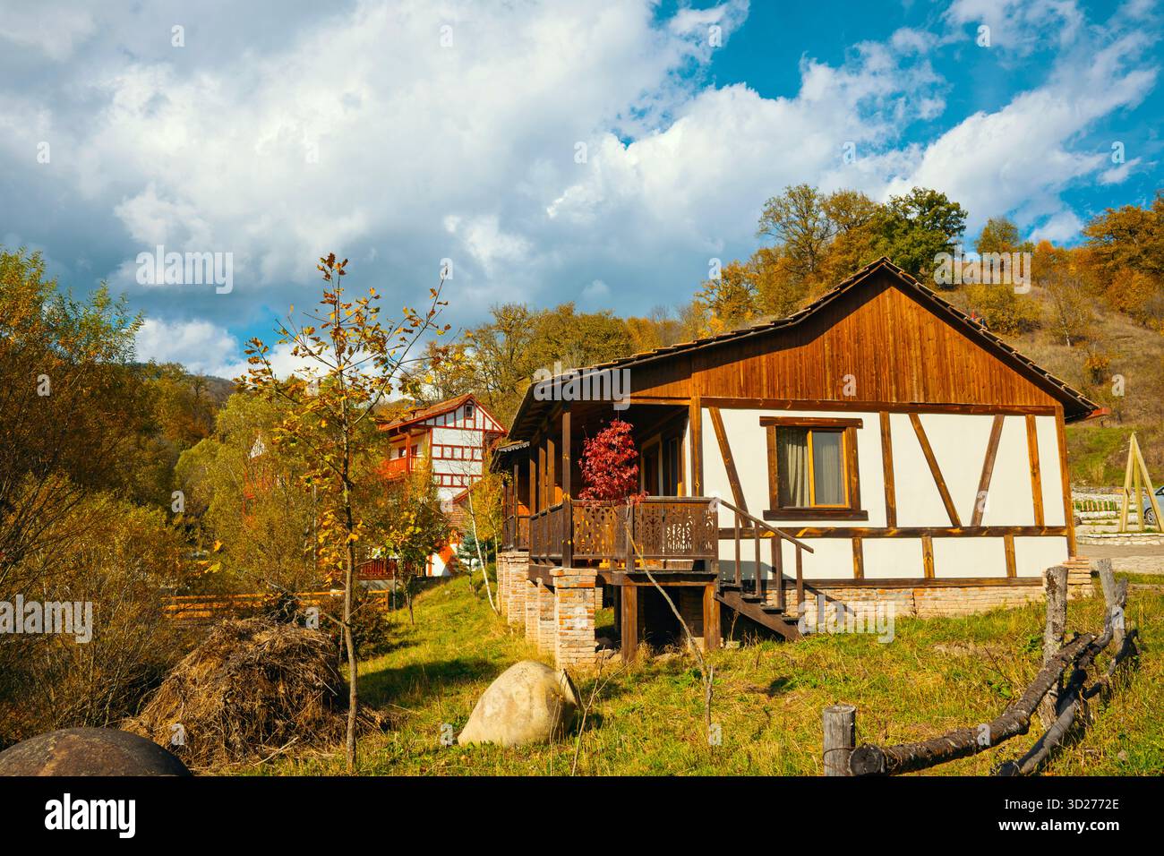 Una piccola casa con un portico si trova in un lussureggiante campo verde. La casa è circondata da alberi e ha un muro di pietra. Il cielo è nuvoloso, ma il sole è ancora Foto Stock