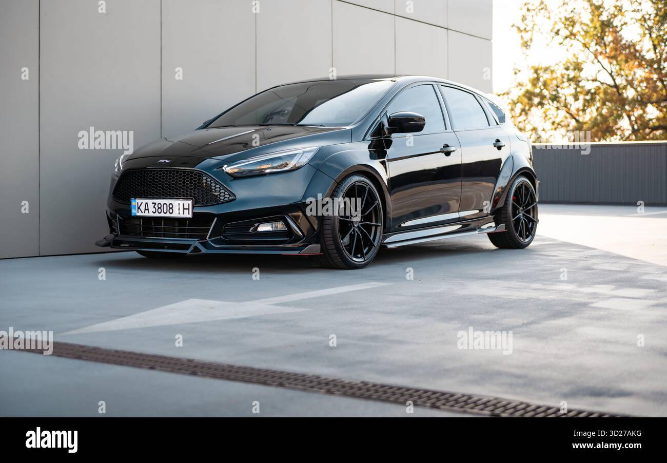 Black Ford Focus nel parcheggio. Vista frontale di tre quarti della vettura berlina modificata vicino al muro beige in una brillante giornata autunnale. Foto Stock