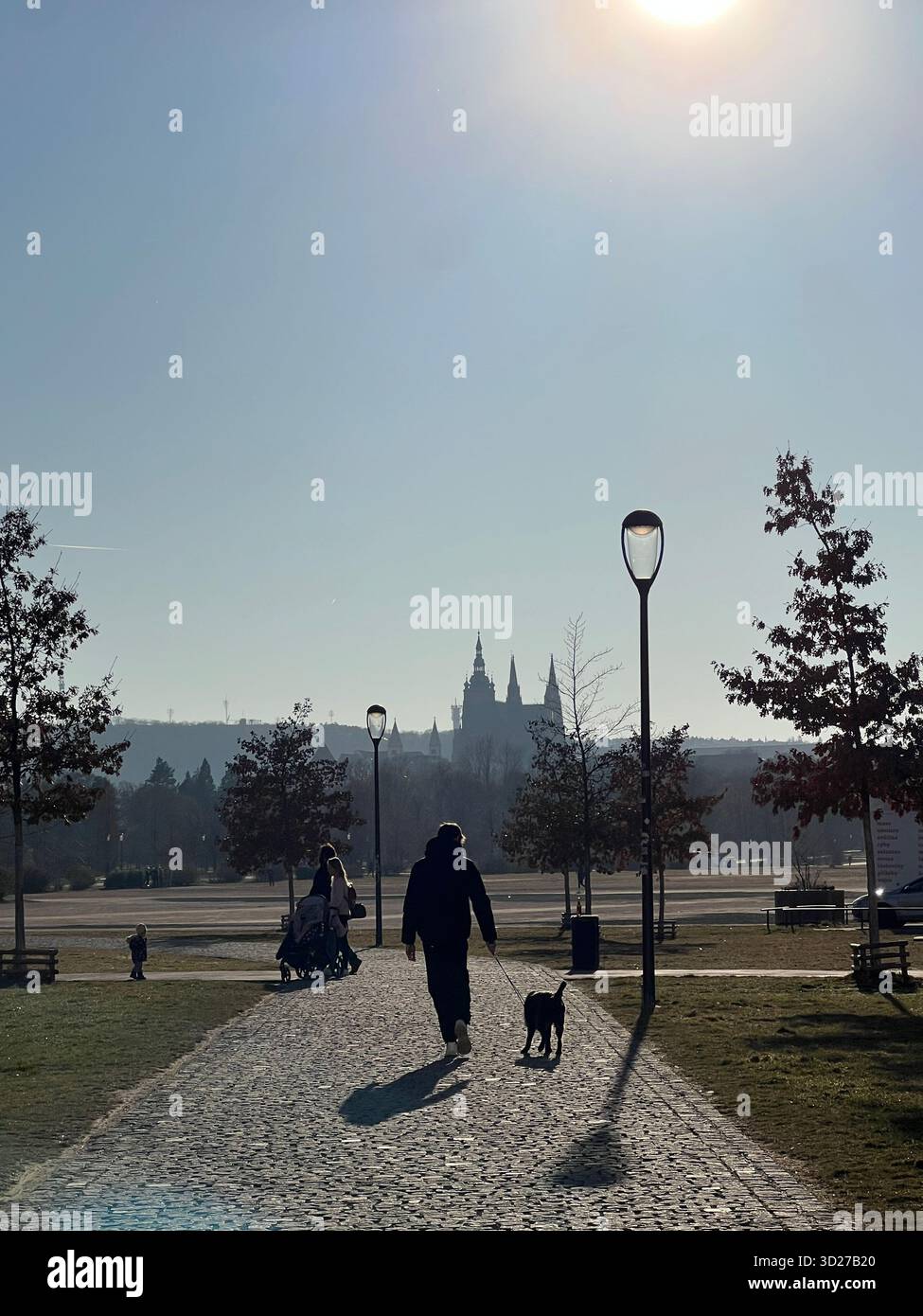 Sagome di persone che camminano in un parco con un cane al tramonto, con il castello di Praga sullo sfondo, Repubblica Ceca. - Immagine stock catturata con smartphone