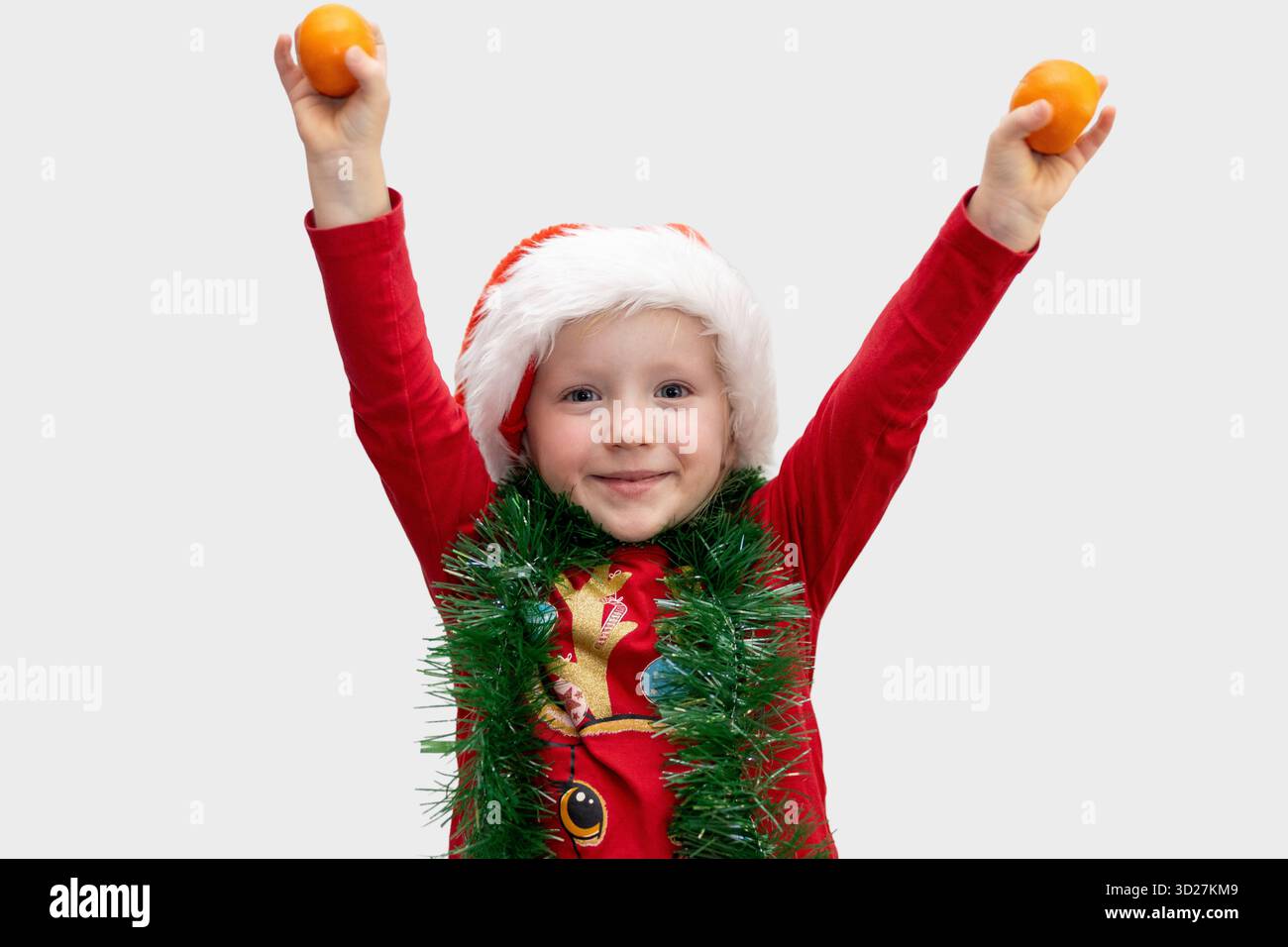 Un bambino allegro vestito con una camicia rossa e il cappello di Babbo Natale tiene le arance in alto nell'aria. La tinsel verde adorna il collo, dando risalto all'emozione per il sacro Foto Stock