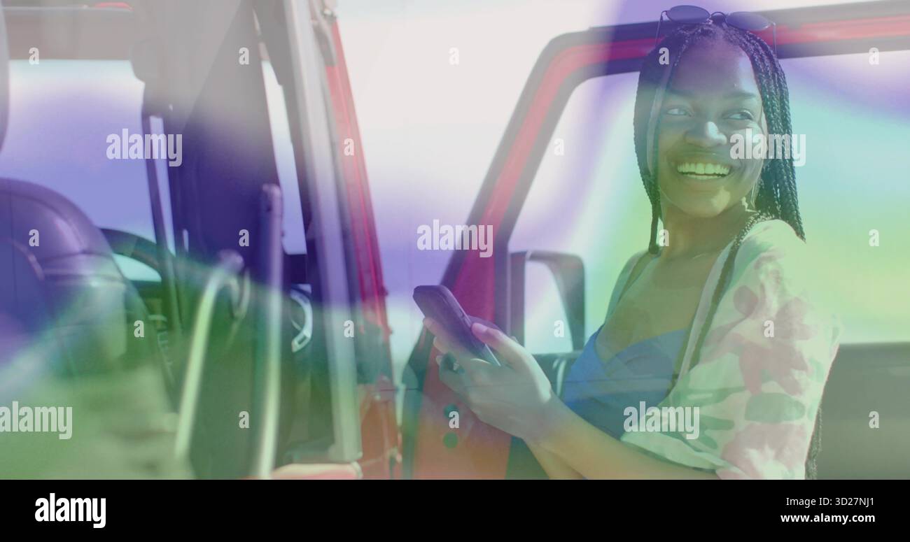 Donna nera sorridente che tiene lo smartphone davanti alla porta rossa del SUV parcheggiata, con volante e cruscotto Foto Stock