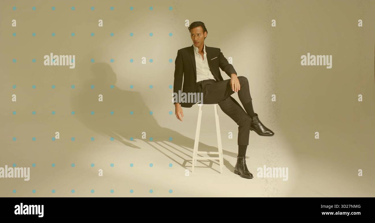 Modello asiatico maschile che indossa una tuta nera in posa sullo sgabello in studio, con sfondo a griglia a punti blu Foto Stock