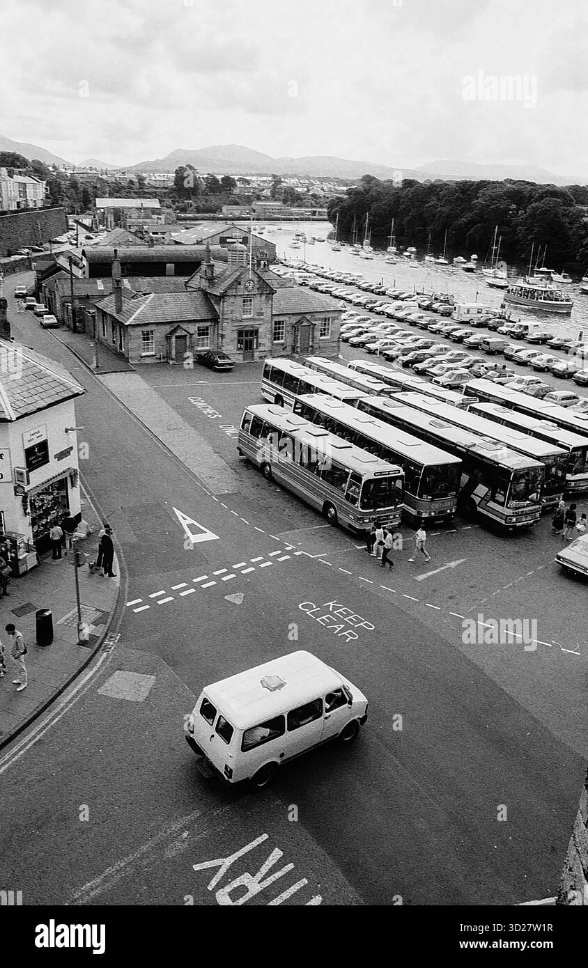 Caernarfon, Galles, una storica città fortificata, offre un'affascinante miscela di architettura medievale e moderni servizi costieri. Questa fotografia cattura una scena di strada trafficata vicino al castello di Caernarfon, mostrando il fulcro dei trasporti della città e la vicinanza allo stretto di Menai. L'immagine evidenzia il collegamento della città con il mare e il suo ruolo di punto di transito vitale all'interno del pittoresco paesaggio gallese. Foto Stock