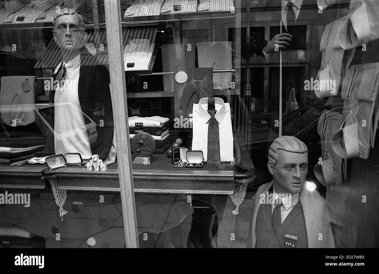 Un chiaro ritratto monocromatico cattura l'eleganza tranquilla di una vetrina di Londra. Due manichini, vestiti impeccabilmente con abiti classici, rimangono congelati in un momento di osservazione silenziosa. La scena, incorniciata dagli interni scuri e strutturati del negozio, evoca un senso di stile senza tempo e la sobria bellezza che si trova nel paesaggio del commercio al dettaglio della città. Uno sguardo nel cuore del quartiere della moda di Londra. Foto Stock