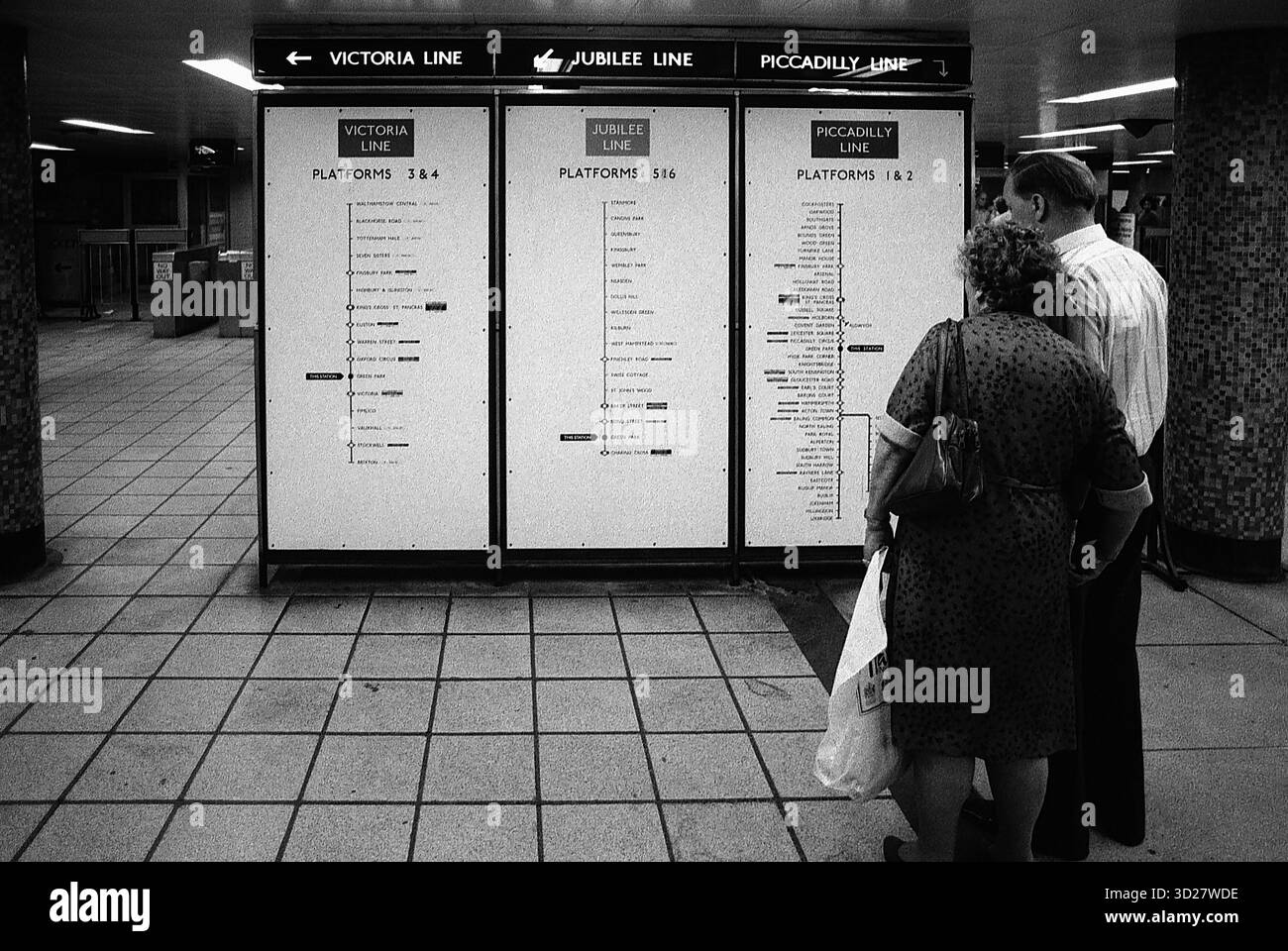 Un momento tranquillo nel mezzo del traffico della metropolitana di Londra. Due personaggi si fermano per consultare la segnaletica della stazione, un'impressionante immagine in bianco e nero che cattura l'essenza di questo storico sistema di trasporto. Foto Stock