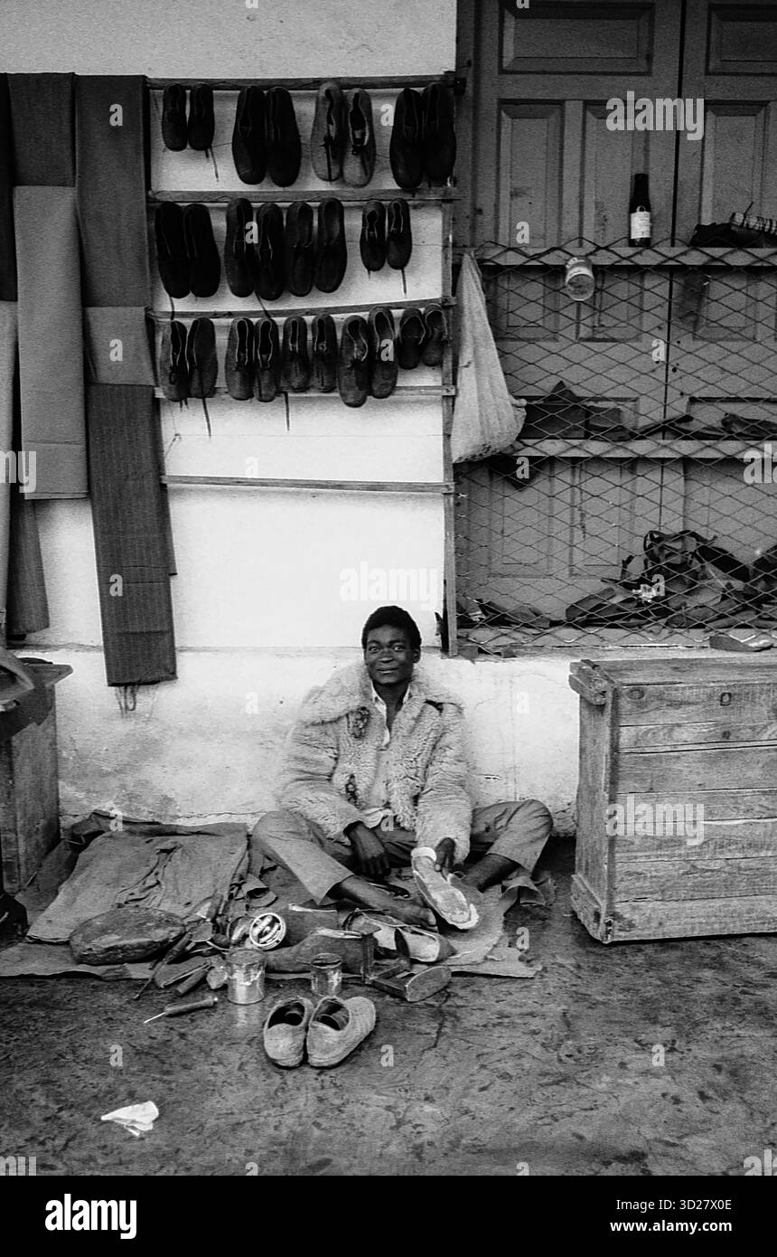 Makuyuni, Tanzania - Un momento tranquillo in una piccola città tanzaniana. Questo ritratto cattura un artigiano locale seduto in mezzo a una collezione di scarpe, che riflette la vita quotidiana e l'artigianato di questa remota regione. L'immagine in bianco e nero accentua le texture dell'ambiente circostante e la presenza dell'individuo, offrendo uno sguardo al cuore della cultura tanzaniana. Foto Stock