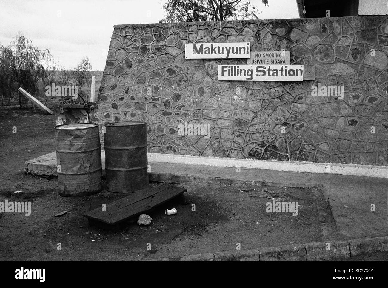 Un cartello con le intemperie che segna la stazione di rifornimento Makuyuni in Tanzania. La stazione si trova in un ambiente rurale, con un muro di pietra dietro il cartello e una collezione di botti nelle vicinanze. Il cartello recita 'Makuyuni' e 'vietato fumare'. E' uno sguardo sulla vita quotidiana in questo angolo remoto della Tanzania, dove il paesaggio e le infrastrutture riflettono un'esistenza semplice ma resiliente. Foto Stock