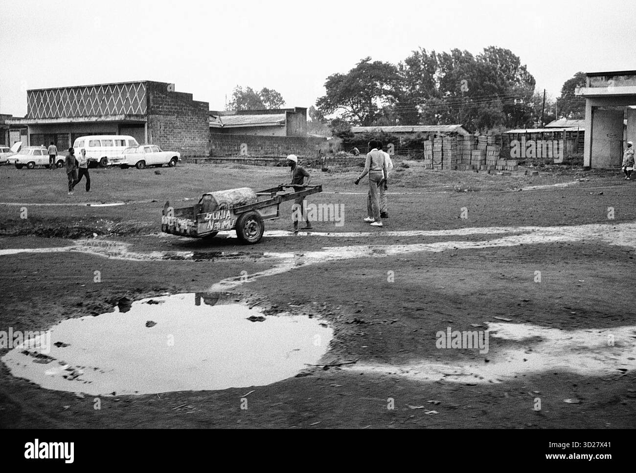 Tanzania: Un momento della vita quotidiana. Questa suggestiva immagine in bianco e nero cattura una scena tranquilla in una città della Tanzania. Un uomo spinge un piccolo carro attraverso una piazza polverosa, mentre altri osservano dall'ombra di un edificio. La scena offre uno sguardo ai ritmi quotidiani della vita in questa nazione dell'Africa orientale, rivelando la semplice bellezza che si trova nei momenti ordinari. Foto Stock