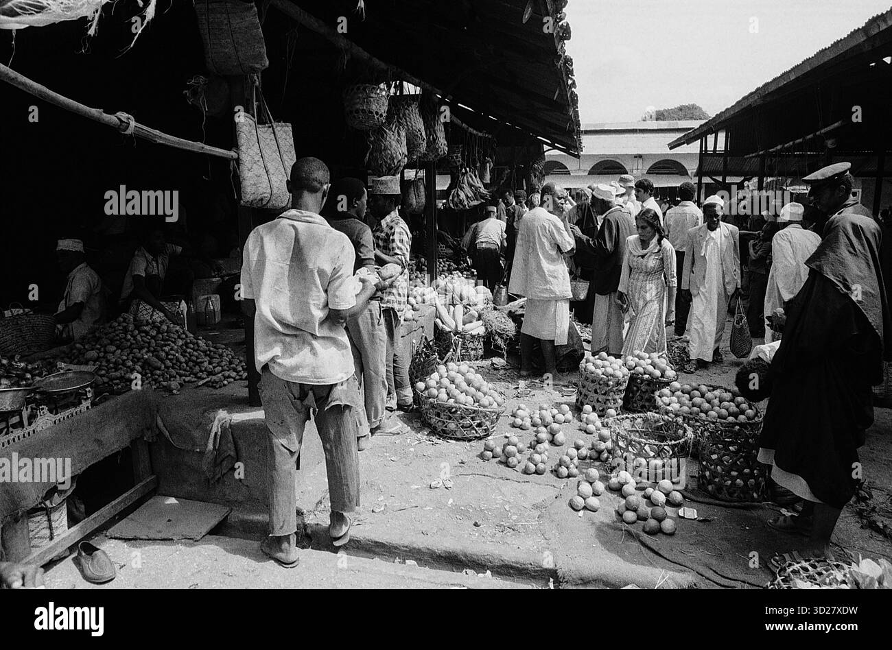 Scena del mercato di Zanzibar: Una fotografia in bianco e nero cattura una vivace scena del mercato a Zanzibar, Tanzania. Gli individui sono presenti in mezzo a una mostra di banane e altri prodotti. L'immagine mostra un tradizionale ambiente di mercato, offrendo uno sguardo al commercio locale e alla vita quotidiana. Foto Stock