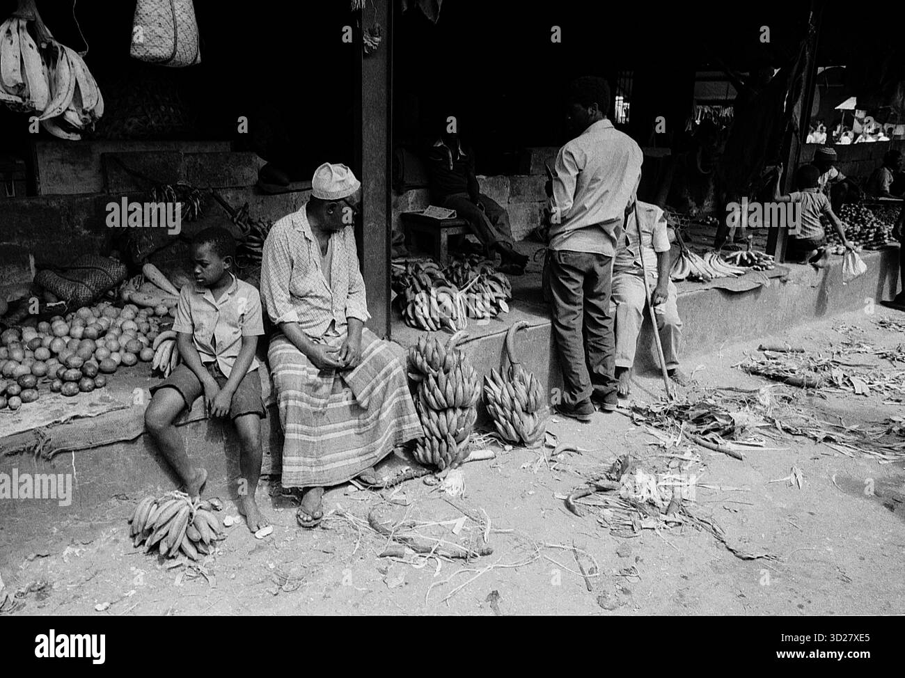 Scena del mercato di Zanzibar: Una fotografia in bianco e nero cattura una vivace scena del mercato a Zanzibar, Tanzania. Gli individui sono presenti in mezzo a una mostra di banane e altri prodotti. L'immagine mostra un tradizionale ambiente di mercato, offrendo uno sguardo al commercio locale e alla vita quotidiana. Foto Stock